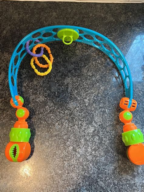 Illustration des Oball Aktivitätsbogens Flex 'n Go, der an einem Kinderwagen befestigt ist, mit den verschiedenen Spielzeugen (Rassel, Affe, Oball).