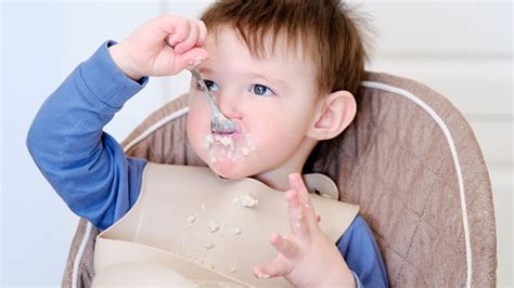 Kleinkind sitzt in einem Hochstuhl und isst Brei mit den Händen, wobei einige Breireste auf dem Lätzchen und dem Tisch sind.